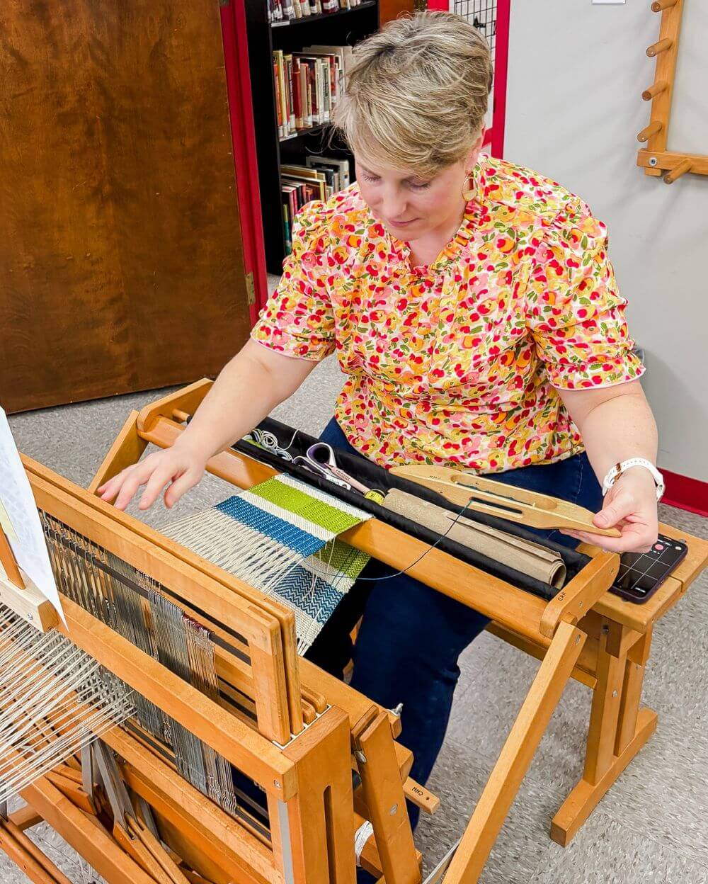 a blonde woman wearing a floral shirt weaves on a floor loom