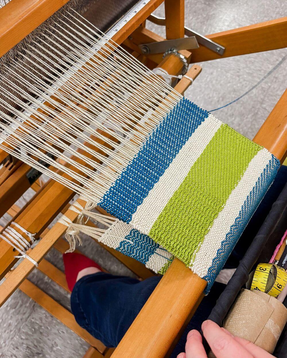 a closeup of a green and blue woven piece of fabric on a floor loom